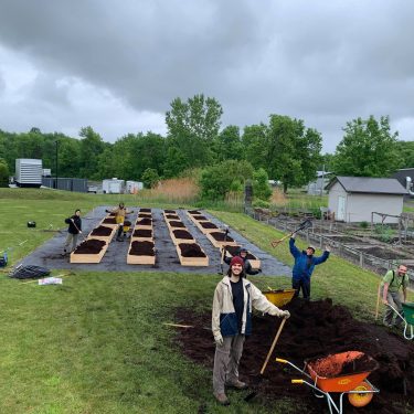 Une équipe de travail devant plusieurs lits de culture remplis de terreau. Ils ont plusieurs outils comme des pelles et des brouettes. Sur le côté, un cabanon derrière un jardin communautaire.