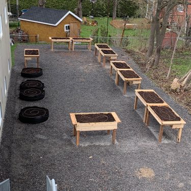 Une dizaine de bacs de culture surélevés et quelques pots de géotextile sont disposés sur une cour de gravier, à l'arrière d'un bâtiment.