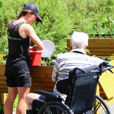 Une horticultrice, debout, et un homme âgé, assis dans une chaise roulante, sont vus de dos alors qu'ils entretiennent des plantes dans un bac de culture surélevé.
