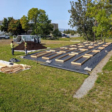 Deux hommes discutent près d'un tas de terre, juste à côté d'une surface recouverte de géotextile où se trouvent 24 cadres de bois.
