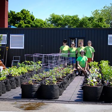 Quatre femmes portant des t-shirts verts identiques se tiennent bras dessus, bras dessous, et un homme portant les mêmes vêtements et accroupi devant elles. L'équipe se tient à l'arrière d'un jardin en pots qui vient d'être planté.