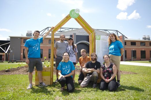 une équipe de 8 personnes devant une arche de bois marquant l'entrée d'un potager. À l'arrière, on aperçoit le Pavillon de la Jeunesse à ExpoCité.
