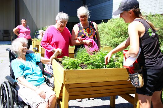 Une horticultrice sourit tout en entretenant le jardin en bac surélevé, alors que trois femmes âgées, dont une en chaise roulante, profitent de la présence du jardin.